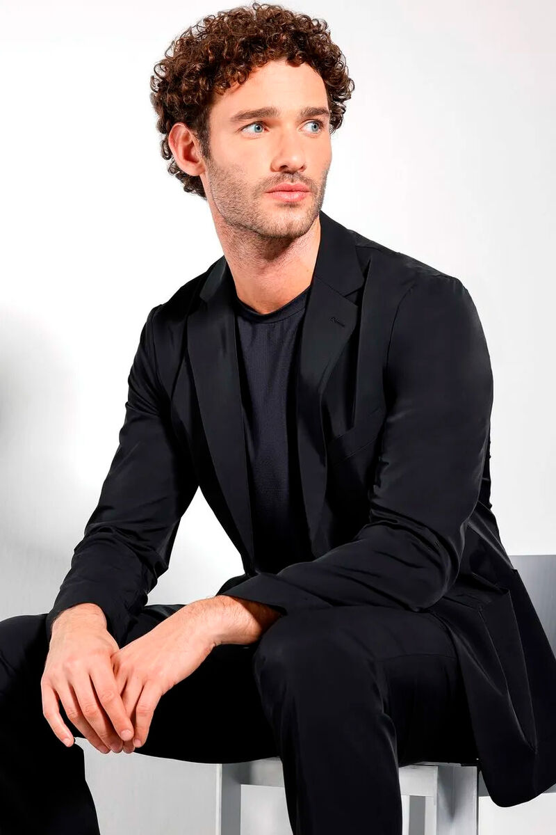 modelo usando uma camiseta preta e um blazer preto sentado em uma cadeira em um fundo branco 