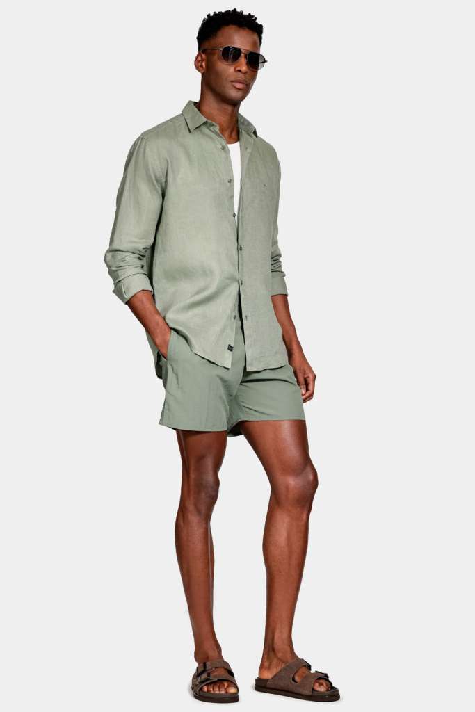 modelo masculino usando uma camiseta branca com uma camisa verde militar por cima aberta e uma bermuda verde junto de um chinelo preto e um fundo branco 