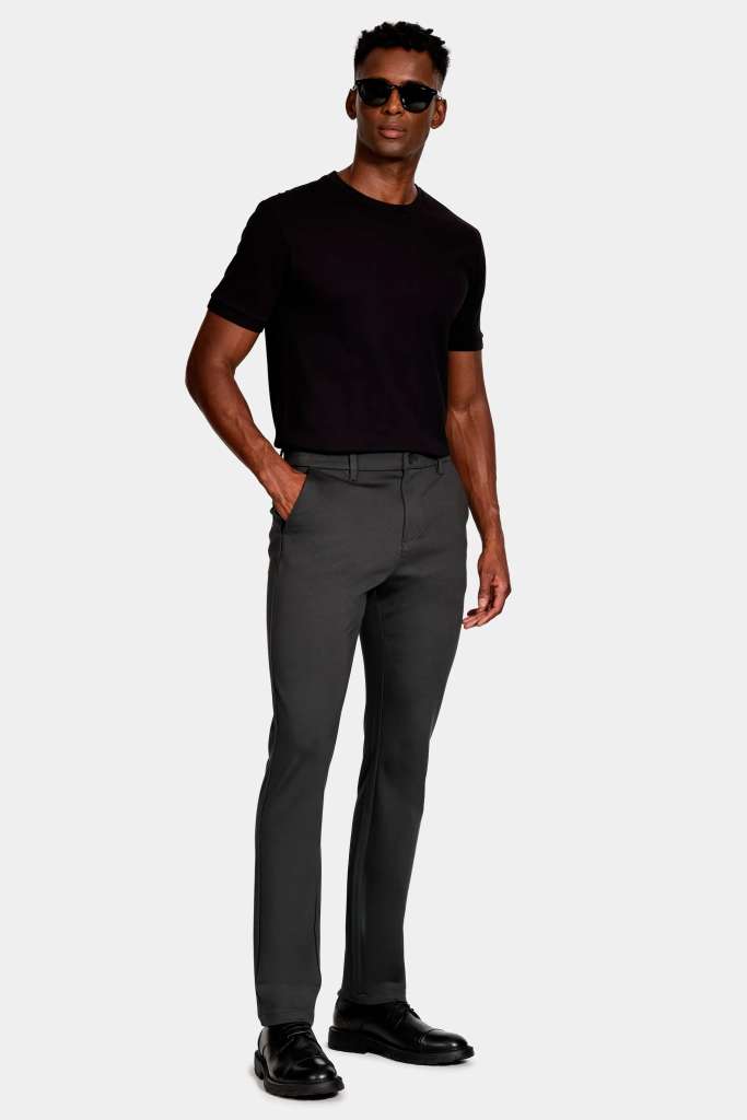 modelo masculino usando uma camiseta preta e uma calça preta, junto de um sapato social preto e oculos escuros em um fundo branco 