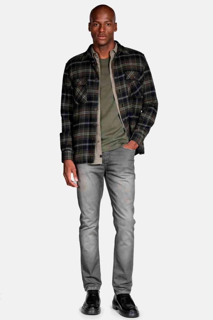 um modelo masculino usando uma camisa preta, uma  camisa xadrez e uma jaqueta junto de uma calça jeans em um fundo branco 