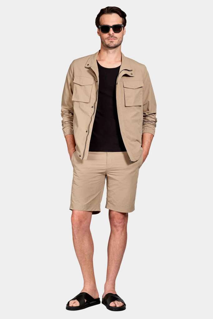 regata slim e bermuda modelo masculino usando uma regata preta com uma camisa sobreposta e uma bermuda bege em um fundo branco