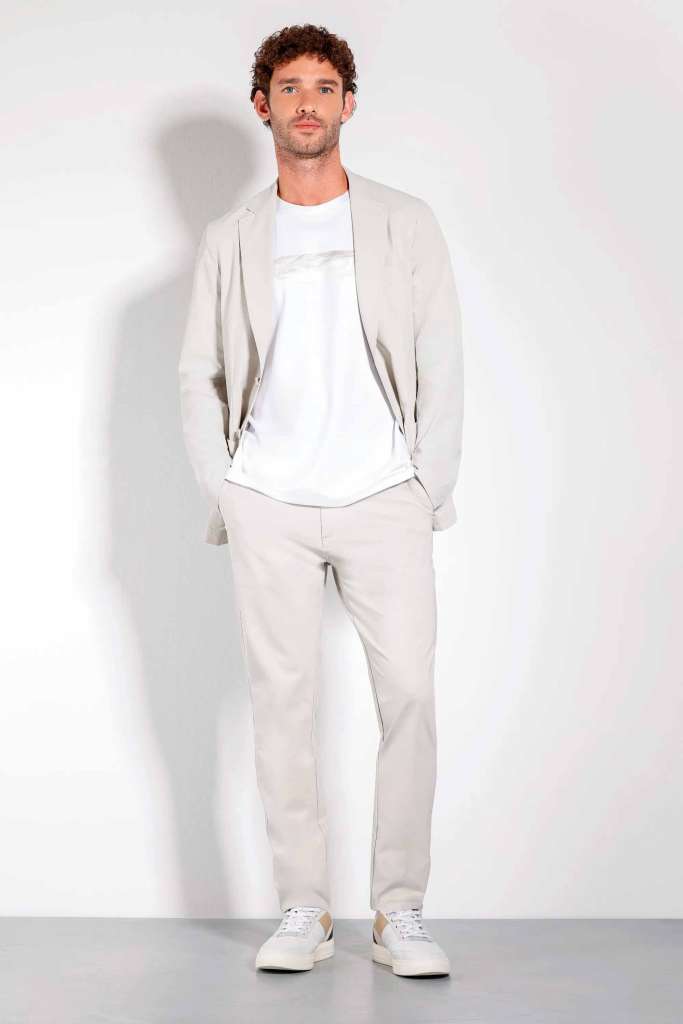 modelo masculino usando uma camisa basica branca e um blazer leve junto de uma calça de alfaiataria em um fundo branco 