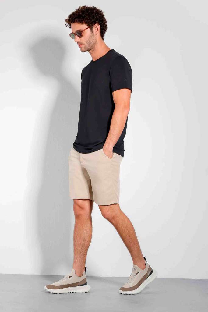 modelo masculino usando uma camiseta preta e uma bermuda bege, junto de tenis e oculos escuros em um fundo branco 