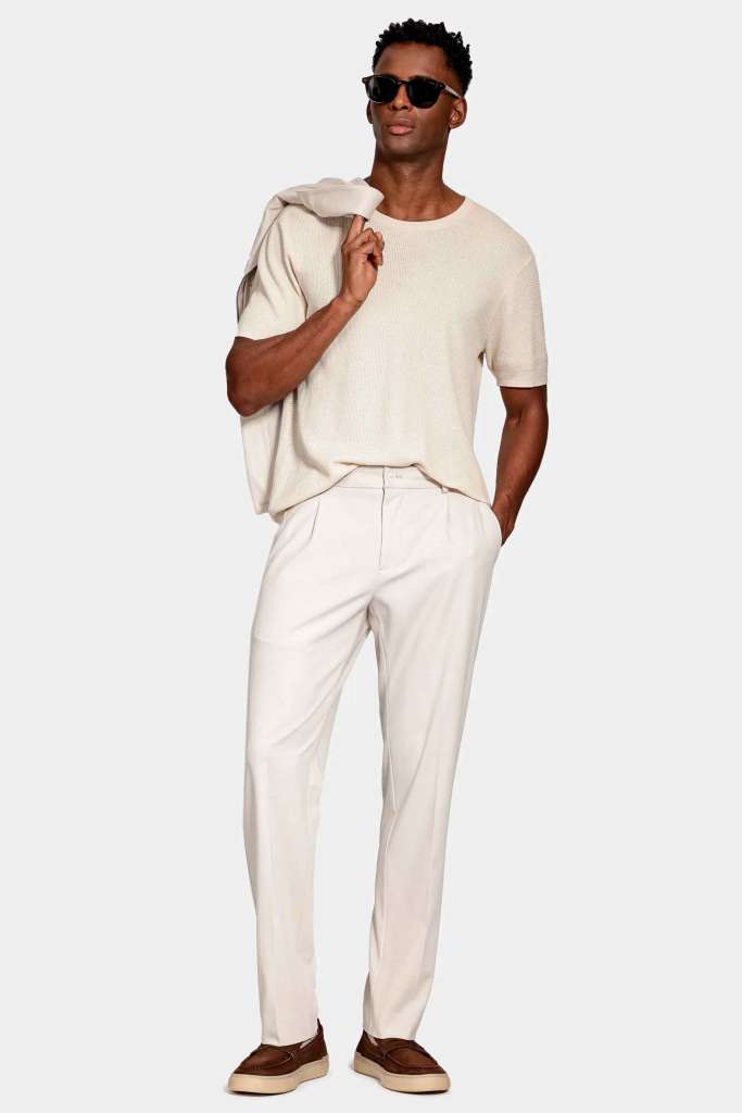modelo masculino usando uma calça alfaiataria bege com uma camiseta bege e um paletó nos ombros em um fundo branco 