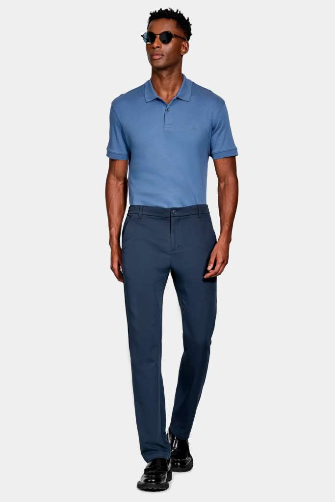 modelo masculino usando uma camiseta polo azul e uma calça preta, junto de oculos escuros e sapatos sociais em um fundo branco 